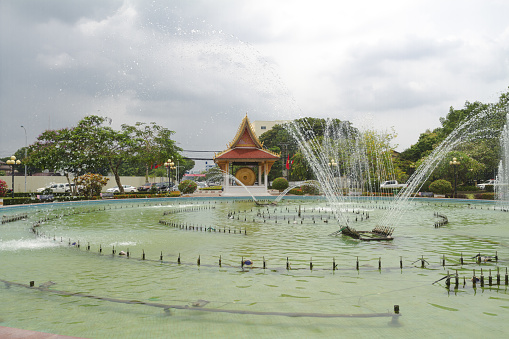 Fountain near Patuxay Monument in Vientiane, part of small inner city Azalea Park. In background are trees, small monument and behind street with traffic