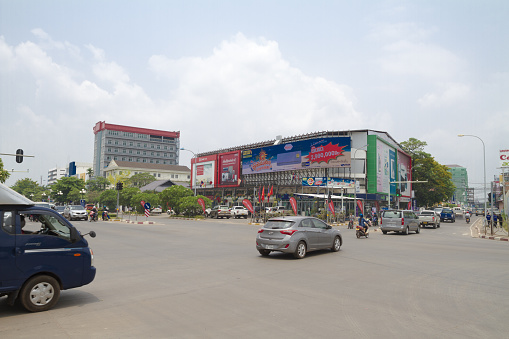 Road intersection in Vientiane with a few traffic and some incidental people in scene. In background are buildings with retail places and stores. At facade are banners and advertising