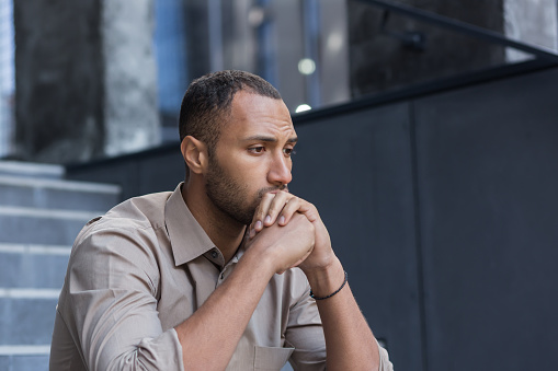 Portrait of a serious and sad hispanic.african american man. He sits sullenly outside on the stairs, holds his head in his hands, looks to the side.