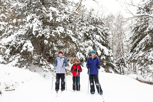 Two sisters and their younger brother stopping to smile for the camera while out cross country skiing in the woods on a winter day.