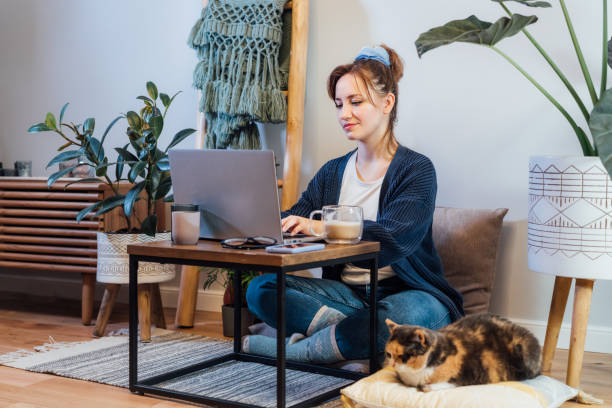 istock Woman using laptop while sitting on floor cushions with sleeping cat pet. Cozy workspace in modern Scandi interior home with green plants. Remote work at home. Online education or entertainment. 1460846878