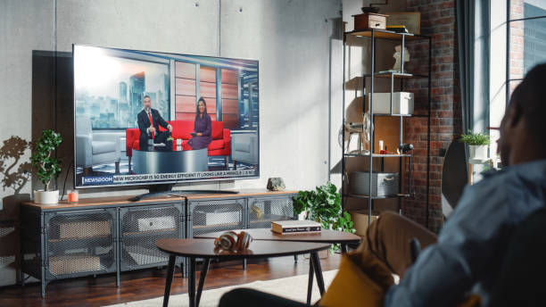 freund und freundin schauen nachrichten im fernsehen, während sie am wochenende zu hause auf einer couch sitzen. zwei moderatoren unterhalten und scherzen auf dem fernsehkanal. gemütliches wohnzimmer mit loft-innenkonzept. - television commercial stock-fotos und bilder