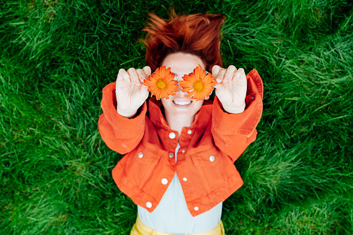 Focus on red orange gerbera Flower glasses in hands of happy smiling redhead woman lying on green grass. Positive Emotion people. Fashion girl in bright colors jeans clothes. Spring, summer mood.