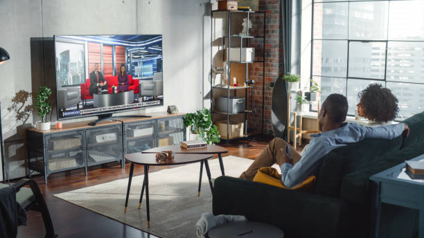 freund und freundin schauen nachrichten im fernsehen, während sie am wochenende zu hause auf einer couch sitzen. zwei moderatoren reden und scherzen im fernsehen. gemütliches wohnzimmer mit loft-innenkonzept. - television commercial stock-fotos und bilder