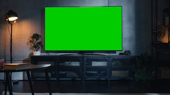 Stylish Loft Apartment Interior with TV Set with Green Screen Mock Up Display Standing on Television Stand. Empty Cozy Living Room of Spacious Flat with Chromakey Placeholder on Monitor.