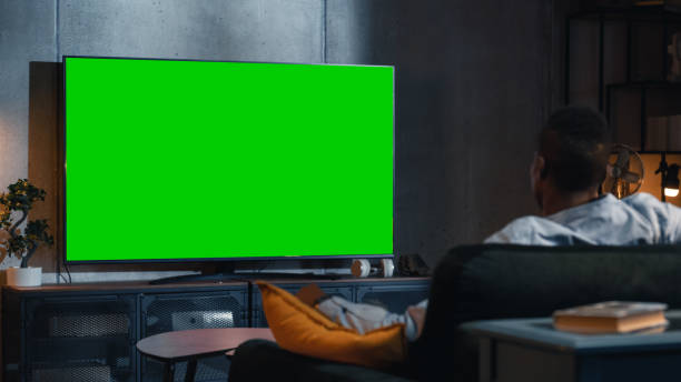 istock Black Man Watches Green Screen Mock-up TV Display with Chromakey While Sitting on a Couch at Home. Relaxed Male Streaming an Evening Movie. Over the Shoulder View. Modern Online Entertainment Concept. 1460848090