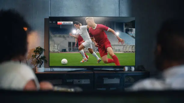 Black Couple Watches Professional Soccer Match on TV, Sitting on a Couch at Home in the Evening. Boyfriend and Girlfriend Football Fans Watch Sports. Back View Out of Focus Close Up Shot at Night.