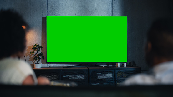 Vista trasera de la pareja negra felizmente casada comiendo palomitas de maíz en el sofá y viendo la televisión con pantalla verde Mock Up Display en la sala de estar del loft. Pareja desenfocada Toma de primer plano por la noche. photo