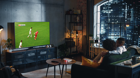 Couple of African American Soccer Fans Relax on a Couch, Watch a Sports Match at Night at Home in Stylish Loft Apartment. Young Man and Woman Enjoying the Game of Favorite Football Club.