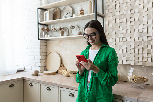 Young amorous teenage girl in glasses and green shirt texting with loved one in red phone at home. Embarrassed, she hides her phone, does not show it, smiles.