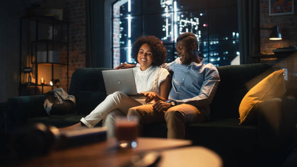 istock Multiracial Couple in Love Use Laptop Computer, while Sitting on the Sofa in Stylish Loft Apartment. Boyfriend and Girlfriend Buy Goods in Internet, Using Social Media Services, Chatting with Friends. 1460849740