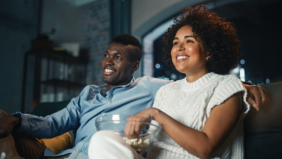 Black Couple Watching Comedy Movie on TV, Eating Popcorn while Sitting on Couch in the Apartment Late at Night. Laughing Boyfriend and Girlfriend Enjoying Funny TV Series at Home. Low Angle Shot.