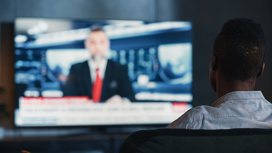 Man Watches Late Night News Show on TV While Sitting on a Couch at Home in the Evening. Presenter Talks on TV. Cozy and Stylish Loft Living Room with Warm Lights. Over the Shoulder Shot.