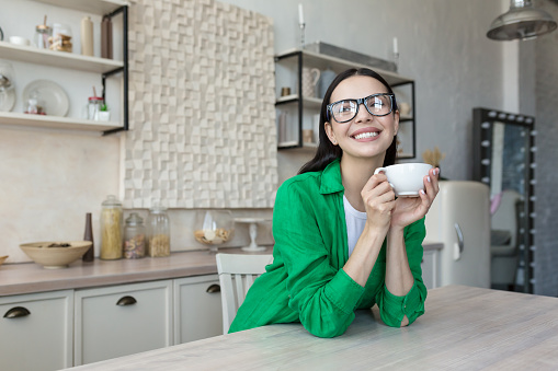 Young beautiful woman, mother drinks coffee, tea alone in the kitchen at home. He holds a cup in his hands, enjoys, rests, smiles at the camera.