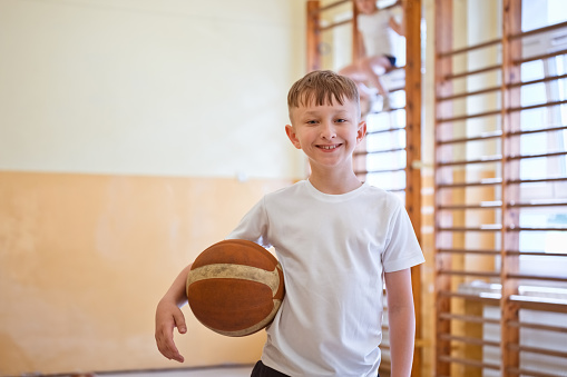 Elementary school boy wearing white t-shirt standing in school gymnasium, holding basketball ball and smiling at camera.