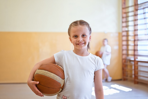 Elementary school girl wearing white t-shirt standing in school gymnasium, holding basketball ball and smiling at camera.