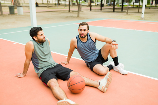 Cheerful attractive men friends resting sitting on the basketball court after playing a one on one game