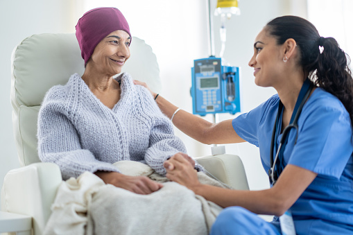 A senior woman battling cancer sits in a high back chair as she talks with her nurse during a Chemotherapy treatment.  The patent is dressed casually in a sweater and has a blanket and headscarf on to keep her warm.