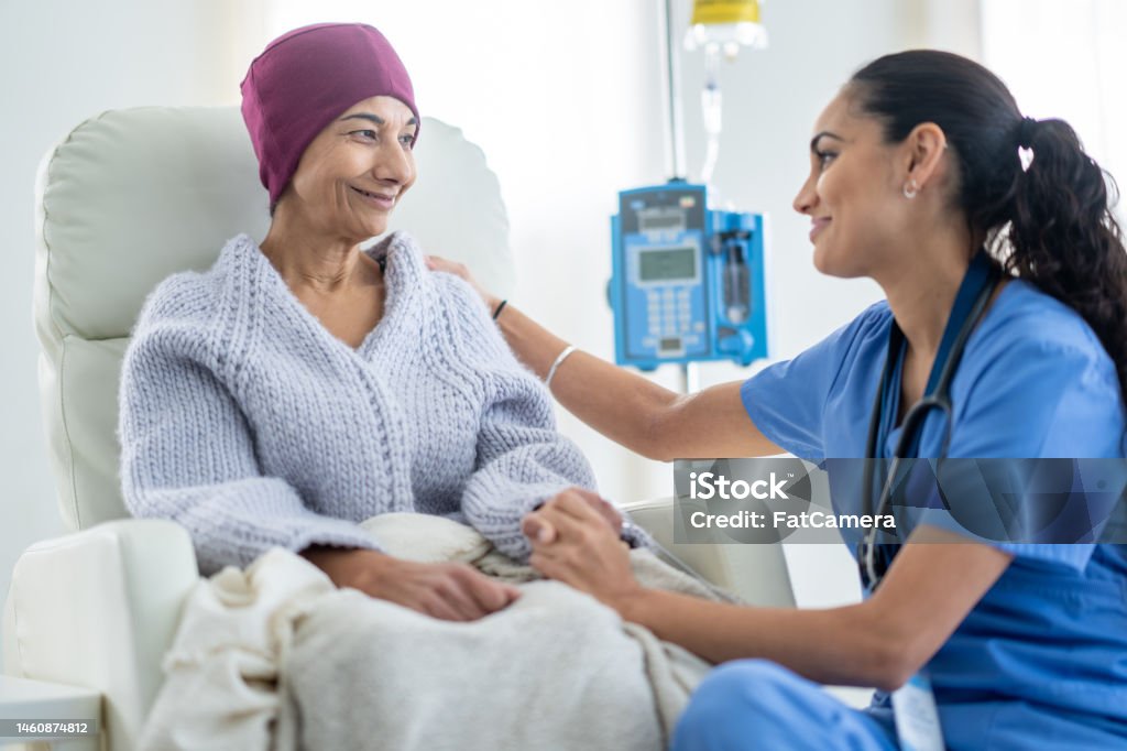 Nurse Talking with an Oncology Patient A senior woman battling cancer sits in a high back chair as she talks with her nurse during a Chemotherapy treatment.  The patent is dressed casually in a sweater and has a blanket and headscarf on to keep her warm. Patient Stock Photo
