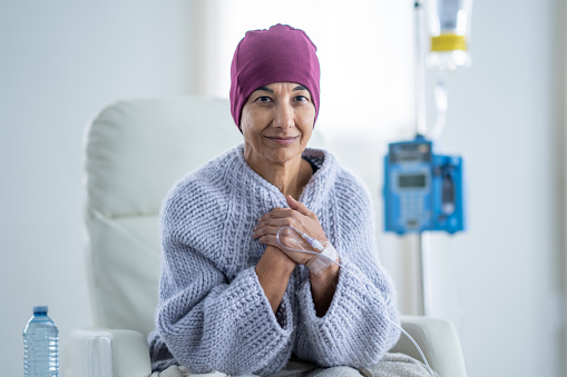 A senior woman of Middle Eastern decent, sits in a high back chair as she receives her Chemotherapy treatment intravenously.  She is dressed comfortably in a sweater and has a blanket and headscarf on to keep her warm.
