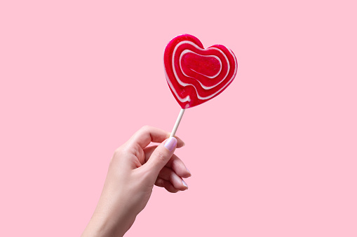 Woman hand holding red heart lollipop on pastel pink background, card for Valentines Day celebration.