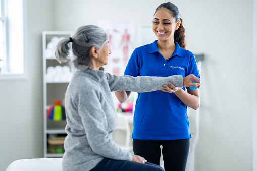 A young female Physiotherapist works her senior patient through a series of stretches as they work on her range of motion and strengthening her arm.  The therapist is dressed professionally as she stands beside the table and supports the woman's arm through each motion.