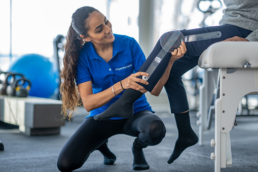 A female Physiotherapist of Middle Eastern decent, sits with a senior patient as she measures her rage of motion and conducts an assessment.  The therapist is dressed professionally as she measures the degrees of flexion in the woman's knee and records it.