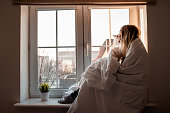 A young woman is sitting near the window wrapped in a blanket and enjoying a sunny frosty winter morning. Cozy winter day at home