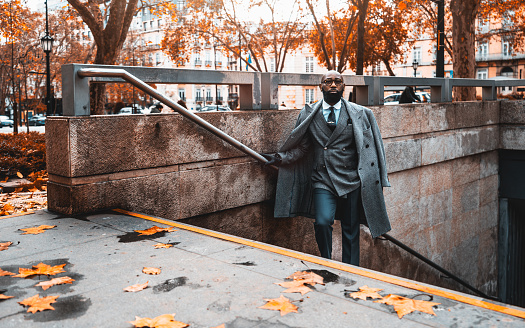Shot of a dandy executive leaning on the metal stairway railing of the stairs of the metro station to the street, he is wearing winter clothes, and the floor is covered with orange-brown leaves