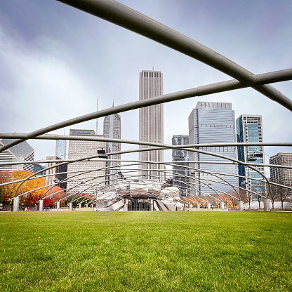 Jay Pritzker Pavilion from a distance, looking north, in Millennium Park, Chicago, on a late fall day with threatening sky. Skyscrapers in background, metal crossbars in forground running through the photo towards the Pavillion across empty green lawn.