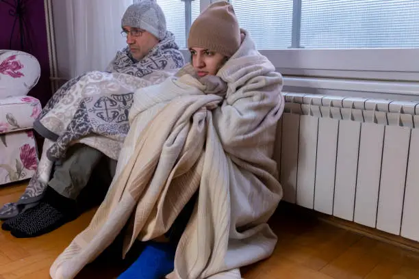 Dad and daughter covered in blankets wearing gloves feel cold using radiator heater to warm up sitting on floor at home in living room during winter