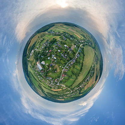 Aerial view from high altitude of little planet earth with small village houses and distant green cultivated agricultural fields with growing crops on bright summer day.