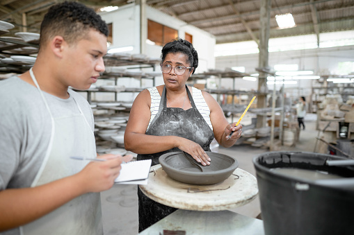 Mature worker showing a young colleague taking notes how to shape a clay bowl in a ceramics manufacturing workshop