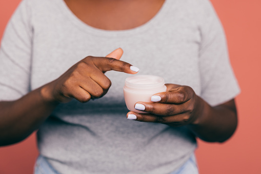 A close up view of an unrecognizable African-American female applying some anti-aging cream on her skin to keep it fresh and smooth. (pink background, skincare concept, copy space)