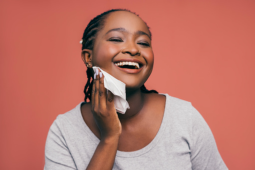 A portrait of a smiling African-American female cleaning her face with a wet wipe while standing against pink background. (studio shot, skincare concept, copy space)