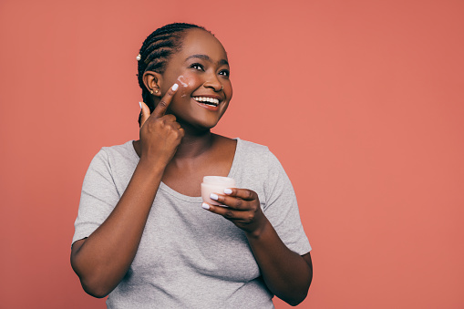 A smiling African-American female applying some anti-aging cream on her skin to keep it fresh and smooth while standing against pink background. (studio shot, skincare concept, copy space)