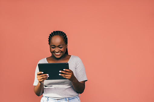 A smiling African-American student laughing while watching something using her tablet. She is standing against pink background. (studio shot, e-learning concept, copy space)
