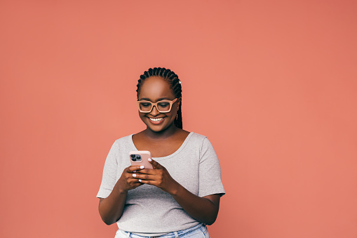 A smiling African-American student watching some educational video on her smartphone while standing against pink background. (studio shot, e-learning concept, copy space)