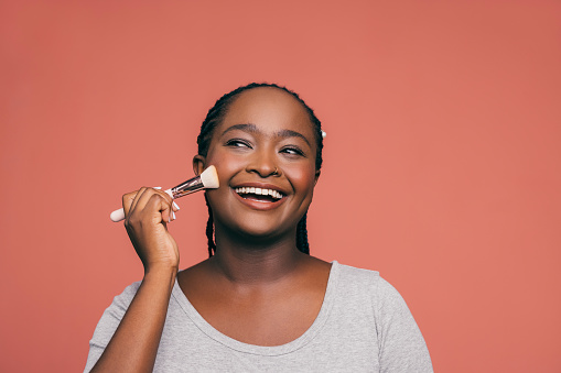 A smiling African-American female applying make up with a brush while standing against pink background. (studio shot, copy space)