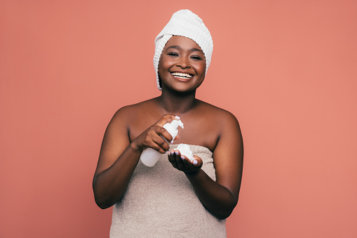 A smiling African-American female applying some foam on her skin to keep it fresh and smooth while standing against pink background. (studio shot, skincare concept, copy space)