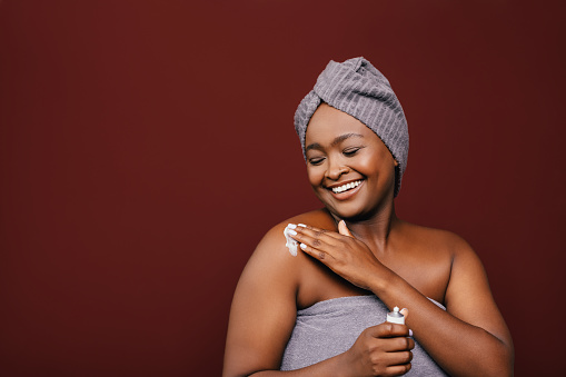 A smiling African-American female applying some anti-aging cream on her skin to keep it fresh and smooth while standing against brown background. (studio shot, skincare concept, copy space)