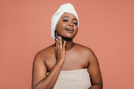 A portrait of a smiling African-American female applying some anti-aging cream on her face to keep it fresh and smooth. She is standing against pink background. (studio shot, skincare concept, copy space)