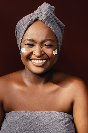 A pretty African-American female laughing while enjoying taking care of her skin by applying some anti-aging cream. She is standing against brown background. (studio shot, skincare concept, copy space)
