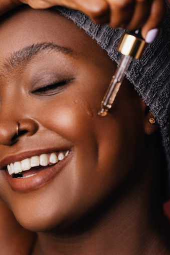A close up view of a smiling African-American female applying some anti-aging serum on her face using a pipette. (brown background, studio shot, skincare concept)
