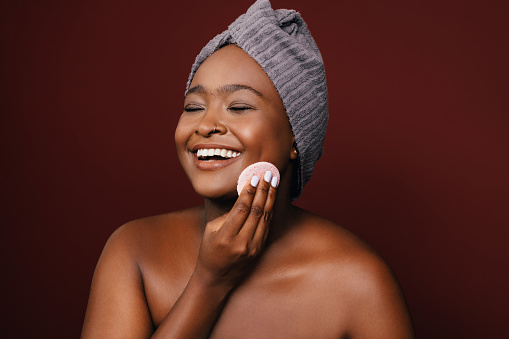 A smiling African-American female taking care of her skin while standing against brown background. (studio shot, copy space)