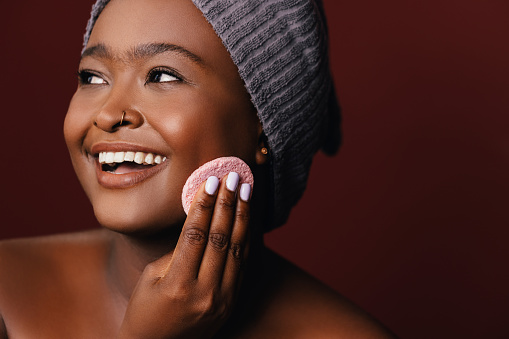 A close up view of a smiling African-American female taking care of her skin. (brown background, studio shot, copy space)