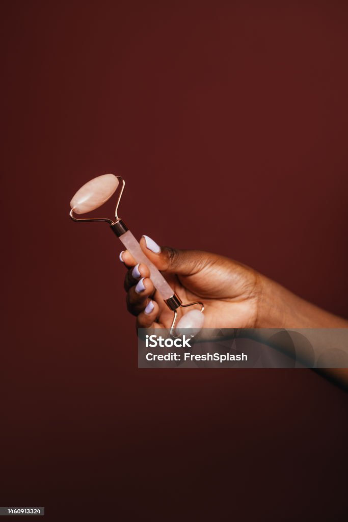 An Unrecognizable Woman Using Jade Roller To Relax A cropped photo of an unrecognizable African-American female holding a jade roller. (brown background, copy space, skincare, self-care concept) Jade Roller Stock Photo