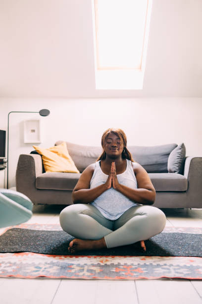 una mujer feliz con sobrepeso haciendo yoga en casa en su esterilla de yoga - young women sitting simple living eastern europe fotografías e imágenes de stock