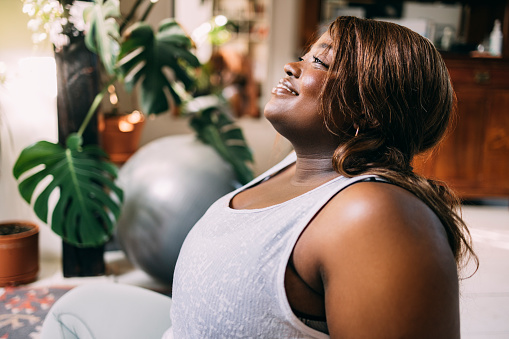 A side view of a smiling overweight African-American female in sports clothes sitting at home and relaxing after doing her daily workout.