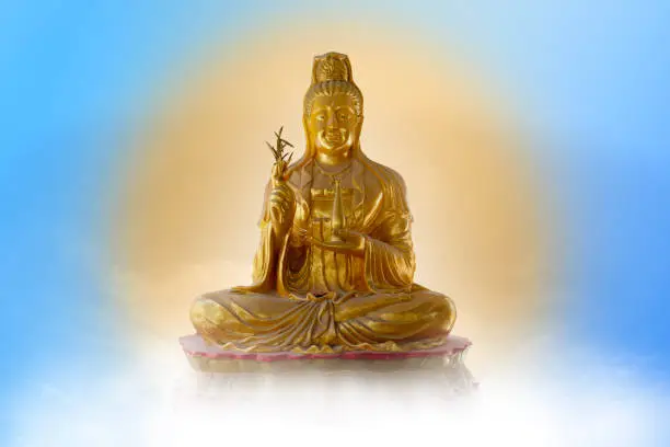 Meditating Golden Guan Yin statue on blue sky background reflecting golden light as if in heaven.